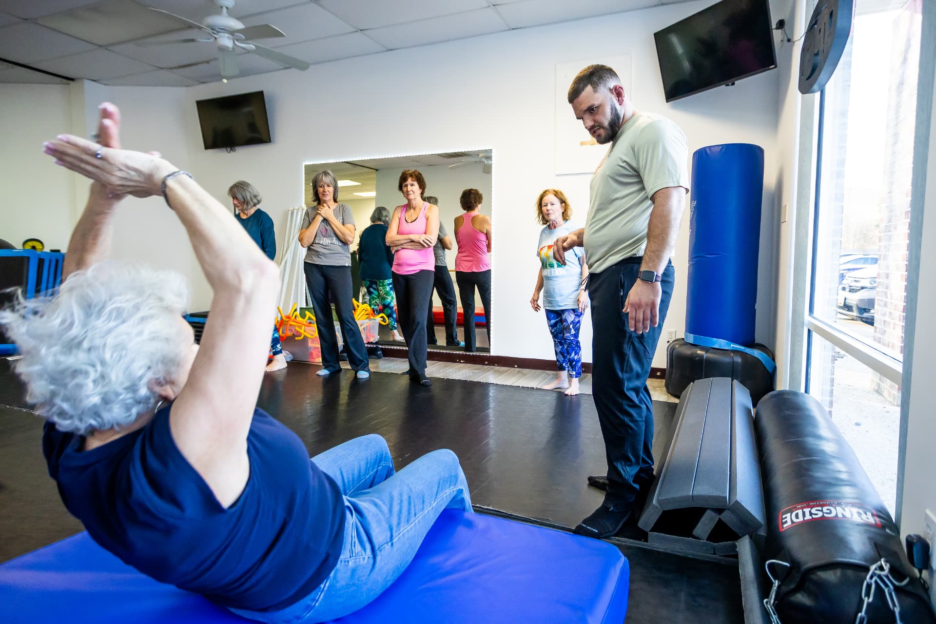 Bryan coaching an older woman through a protective backward movement on a soft mat while classmates watch