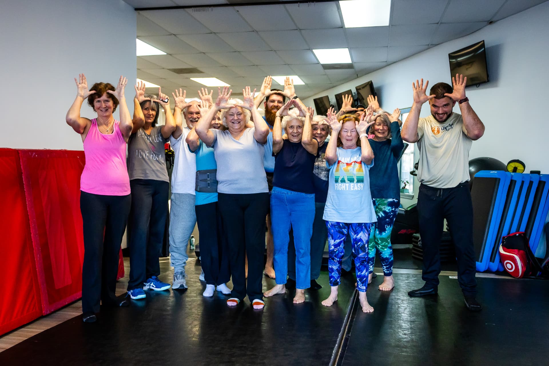 A smiling group of older adults and two Ukemi Health instructors raising hands in a protective guard after class
