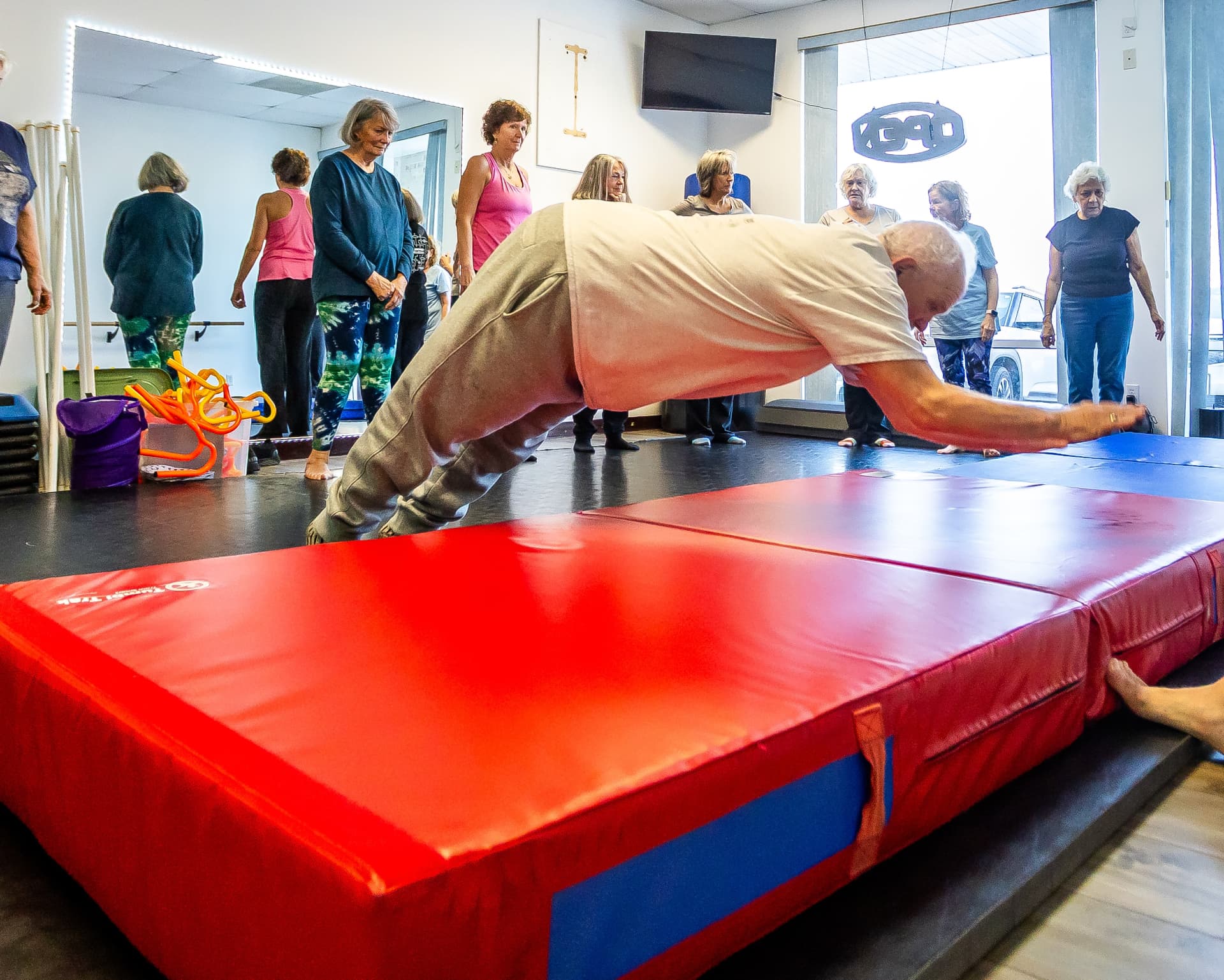 An older man practicing a supported forward movement onto a thick padded mat as classmates look on